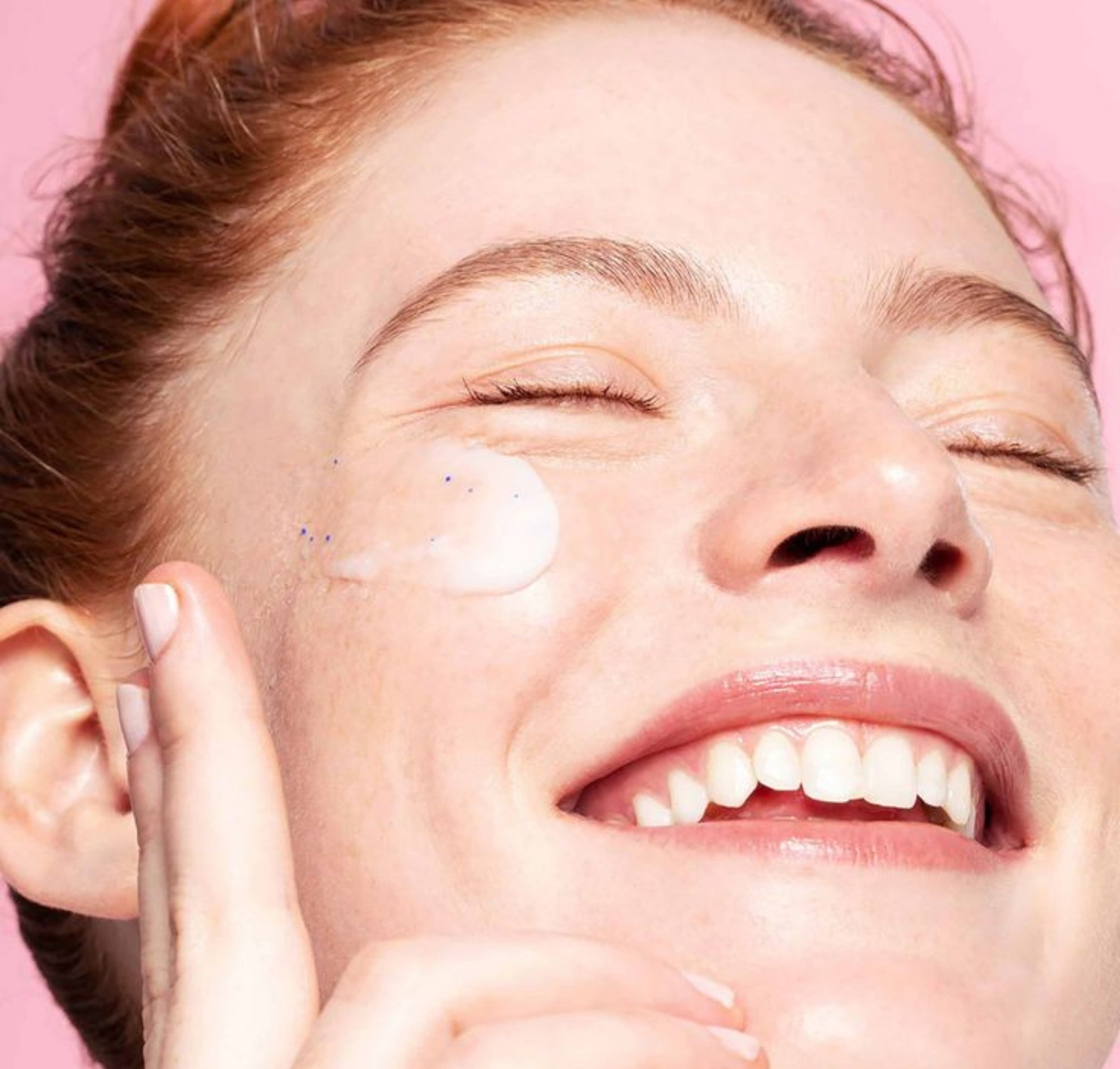 Close-up of a person applying cream to their face with a pink background